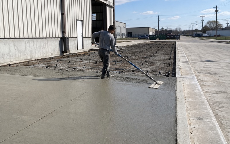 A dowel basket being set at a construction joint in a commercial truck apron in Kyle, the formed pavement running away toward a loading dock.